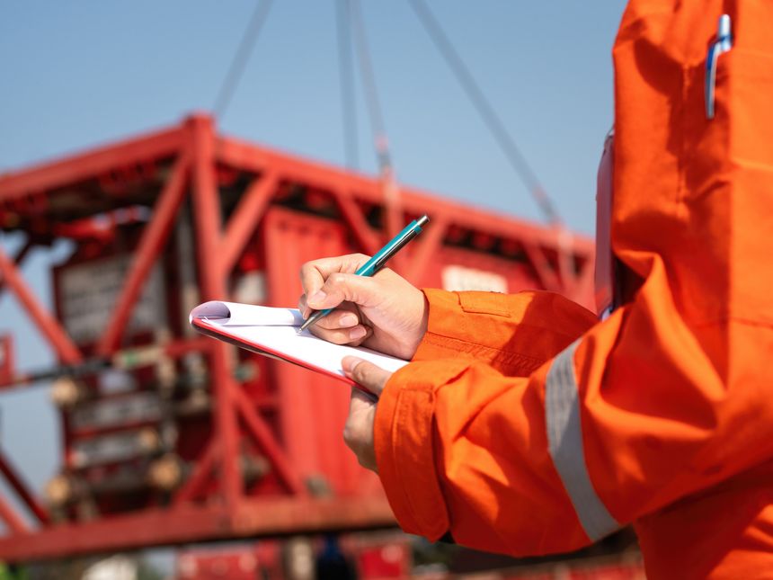 A safety officer is checking on safety checklist form during perform inspection at industrial working site with heavy lifting operation as background. Close-up and selective focus.