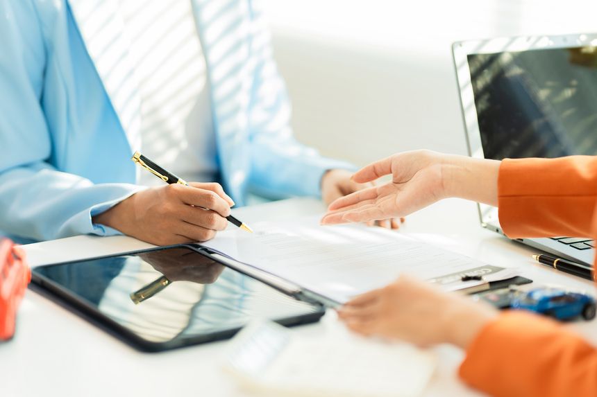 A car dealer is explaining contract terms to a customer inside the office; a close-up shot of a salesperson showing vehicle purchase documents.
