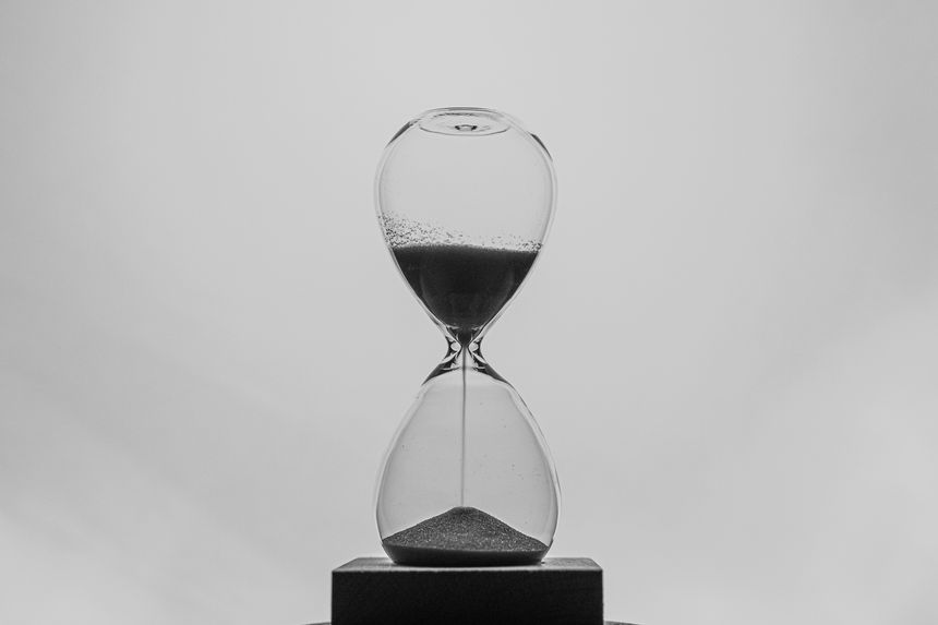 A black and white photograph of an hourglass with sand flowing from the top chamber to the bottom. The minimalist background emphasizes the concept of time and its passing.