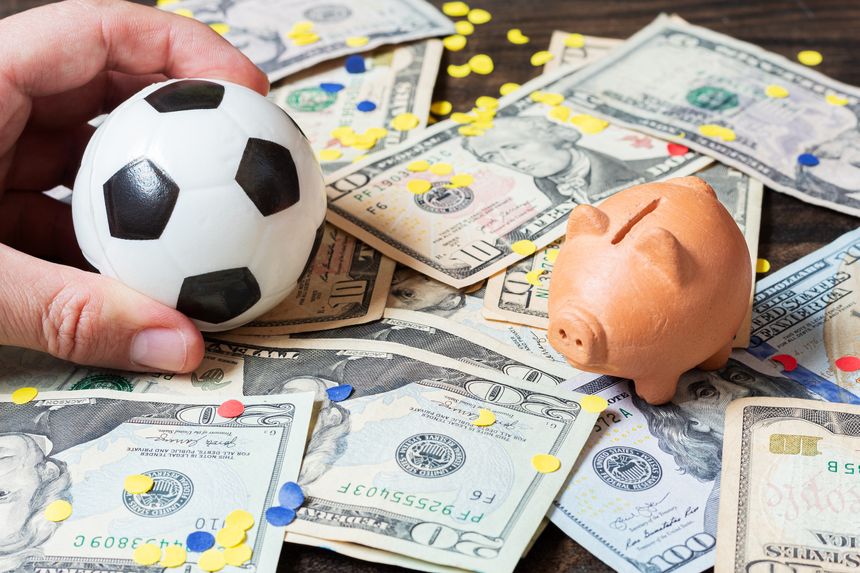 Hand holding a soccer ball on a table covered with dollar bills next to a piggy bank, concept of soccer economics and sports betting