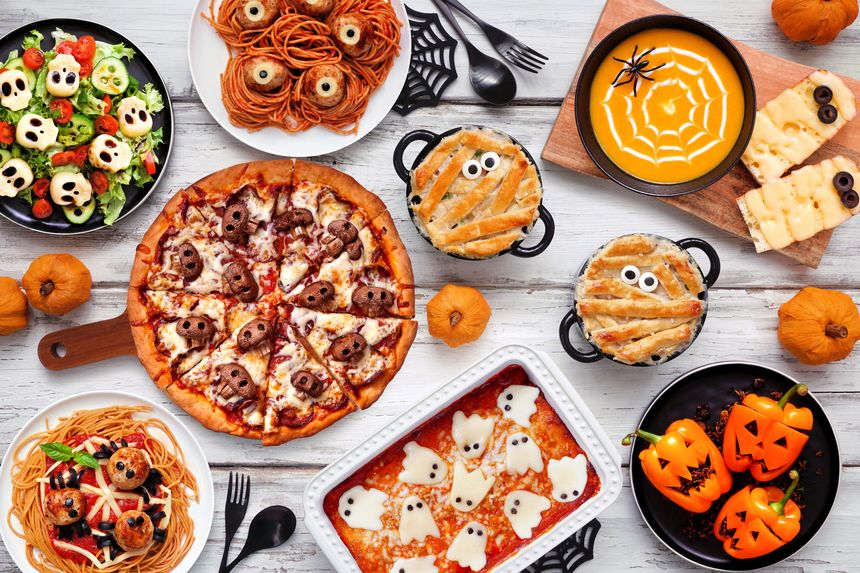 Spooky Halloween dinner party table scene over a white wood background. Top view. Skull pizza, salad, pasta, ghost lasagna, mummy pot pie, pumpkin soup, jack o lantern stuffed peppers.