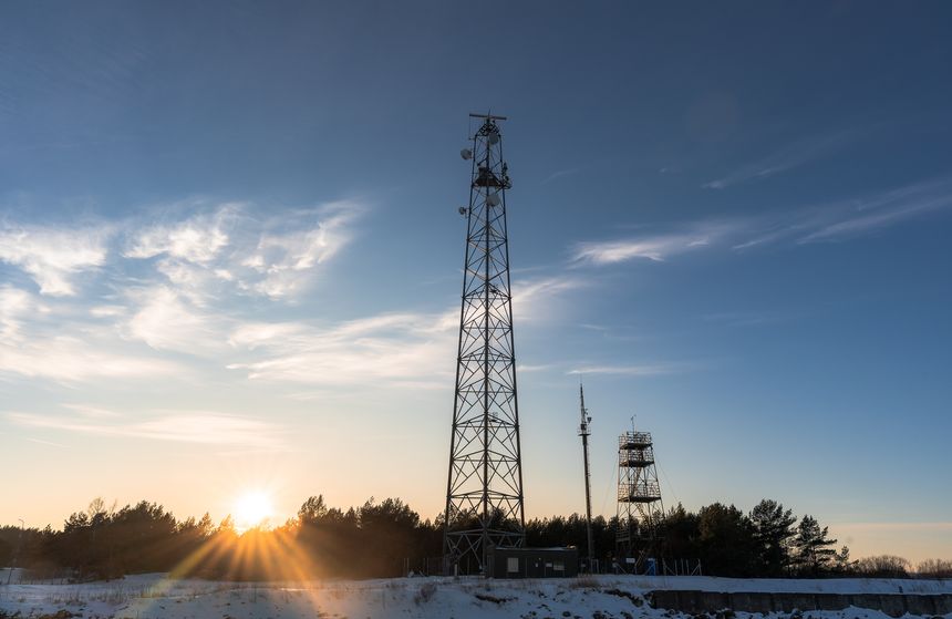 Tall telecommunication tower with radar and antennas against a blue sky at sunset. Winter landscape with snow, forest silhouette, and bright sunburst flare through the trees.