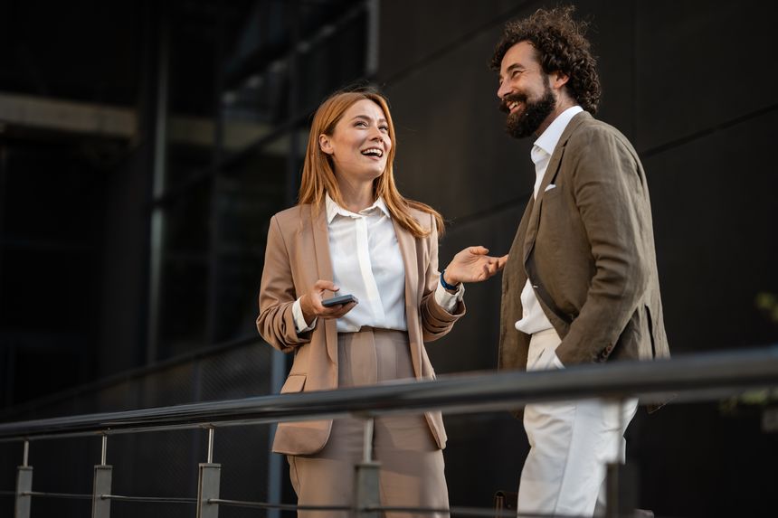 Two cheerful businesspeople engaging in lively conversation and laughter during a break outside their office, enjoying the sunny urban atmosphere