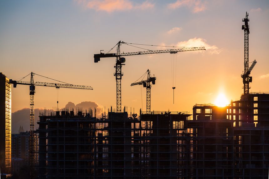 Lots of tower cranes build high-rise residential building in evening time. Steel frame structure in sunlight. Industrial background with sun flare. Yellow sunset sky.