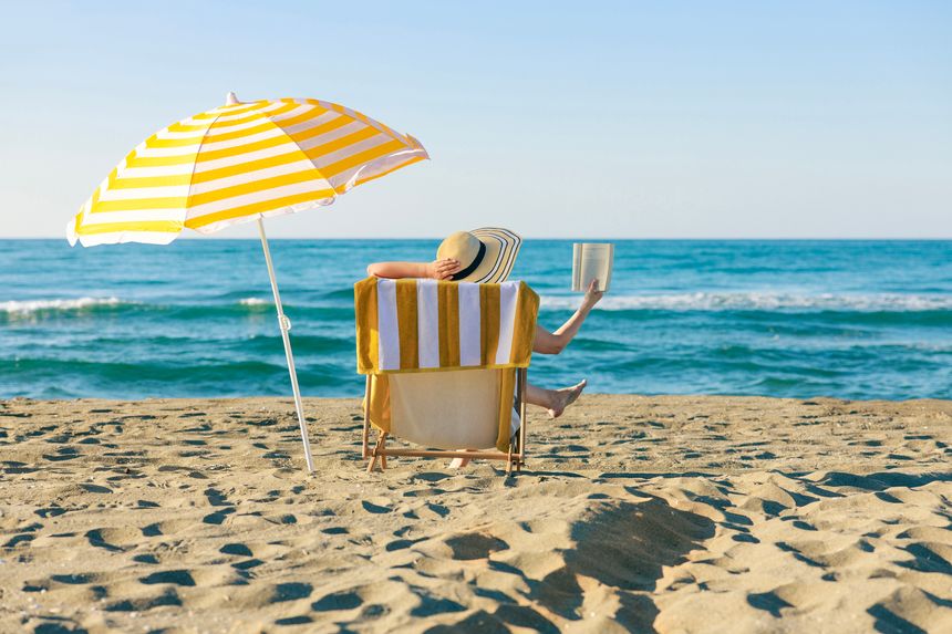 Woman relaxing on a beach chair under a striped umbrella while reading a book by the ocean. Calm summer atmosphere with vacation and leisure concept
