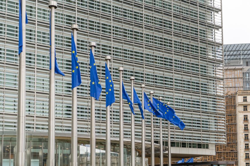 Brussels, Belgium. A row of European Union flags on poles in front of the Berlaymont building, with a modern glass facade and strong linear structure dominating the background.