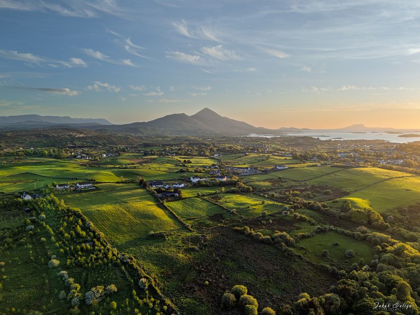 A breathtaking panoramic view of the iconic Croagh Patrickrising from the lush green Irish countryside. Bathed in warm golden light of the setting sun, gentle rolling hills lead the eye towards the majestic peat, creating a scene of tranquility and natural beauty in County Mayo, Ireland.