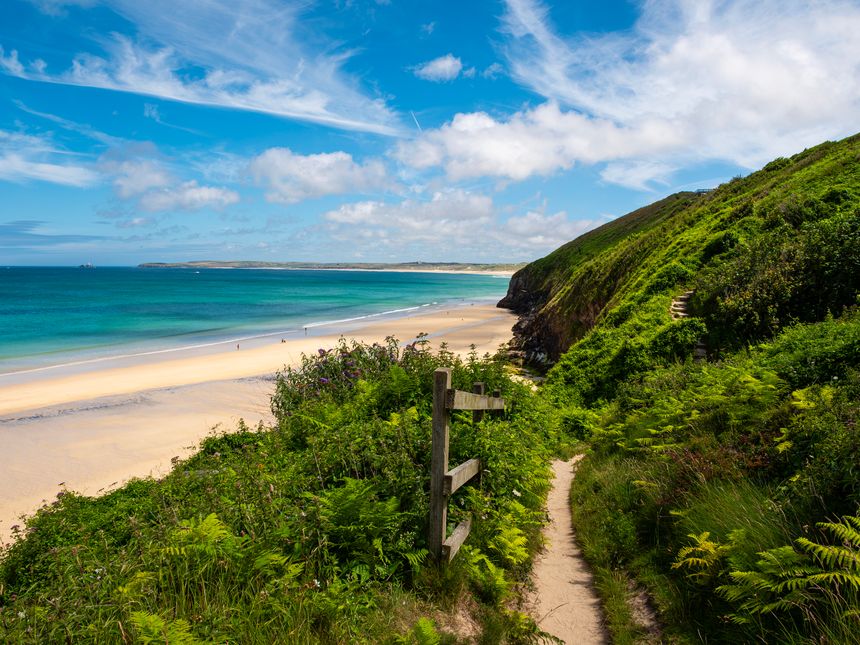 The South West Coast Path, passes Carbis Bay, Cornwall, UK, along scenic views of the bay, sandy beach and coastline. Blue sea, bright sky.