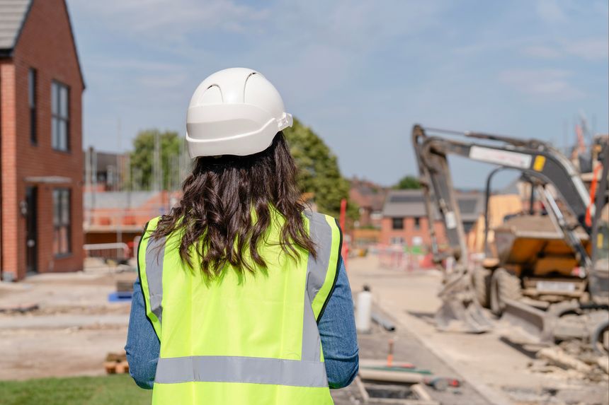 Woman builder engineer in  bright safety jacket and hard hat uses surveying instrument at a construction site surrounded by construction materials and barriers