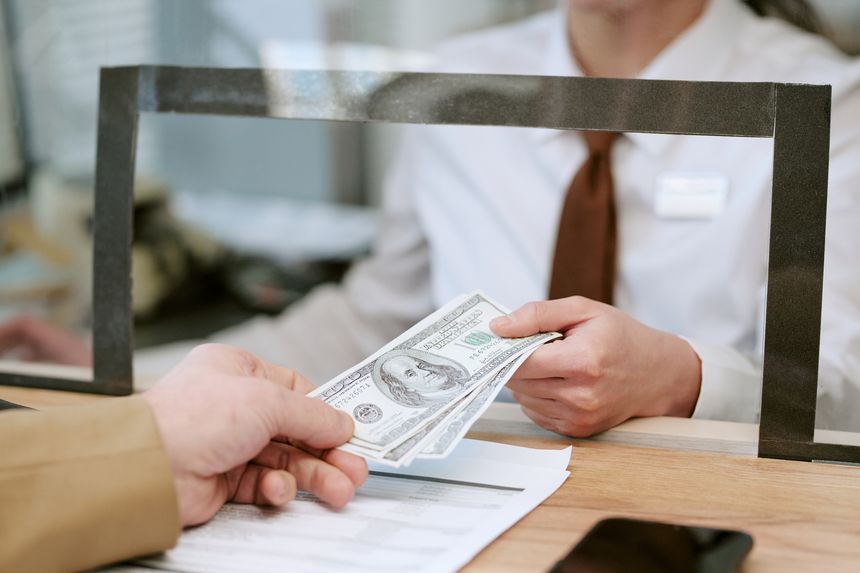 Female bank teller handing cash to Caucasian middle aged man customer through counter window, both interacting during financial transaction at bank branch