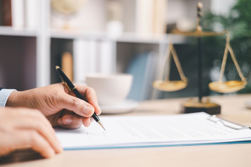 Lawyer in suit signing a contract on clipboard at office desk with justice scales in background, representing legal services, compliance review, agreement approval and documentation.
