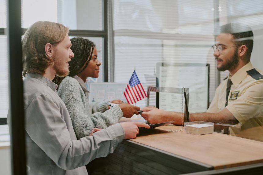 Diverse couple handing passports at immigration office counter with officer verifying documents and flag in background. Multi-ethnic group interacting in official setting