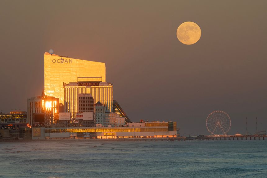 An afternoon view of the Atlantic City skyline with the casinos illuminated by the setting sun and a full moon rising in the background