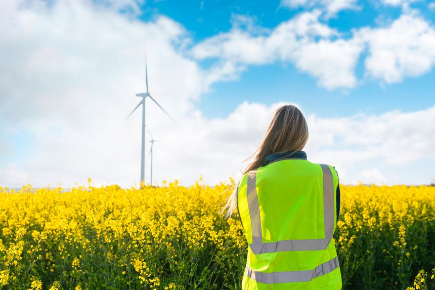 A person wearing a bright vest stands in a field of yellow flowers. Wind turbines are visible in the background on a sunny day with some clouds.