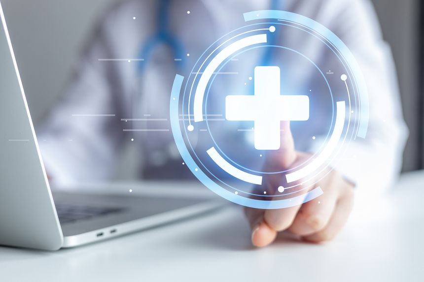 A medical professional points to a digital health symbol on a screen while seated at a desk with a laptop. The office setting shows a focus on technology in healthcare.