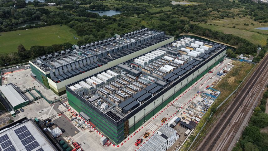 Two large data center buildings stand beside a railway line. Extensive rooftop infrastructure and modular units are visible. Green fields, trees, and lakes surround the complex on a bright sunny day.