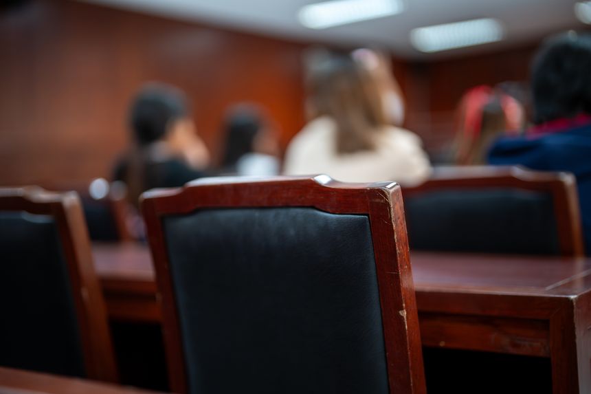 A group of people are sitting in a room with wooden chairs. The chairs are black and the room is dimly lit