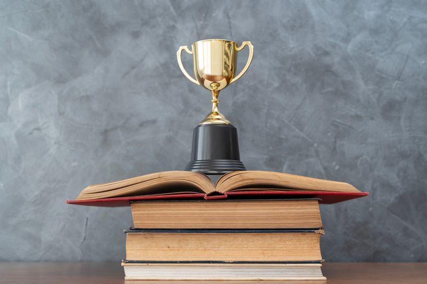 Gold trophy placed above stacked books and an open page, symbolizing academic excellence, learning achievement, student success, and educational recognition.