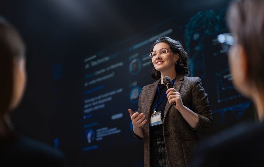 A confident young businesswoman with glasses delivers a compelling speech on stage at a technology conference. Holding a microphone and wearing a name badge, she connects with her audience. The digital screen behind her displays high-tech visuals, emphasizing innovation and leadership. This dynamic scene captures professional ambition, public speaking, empowerment, and the exciting energy of the tech industry. Ideal for illustrating leadership, business growth, industry expertise, and empowerment in modern professional environments.