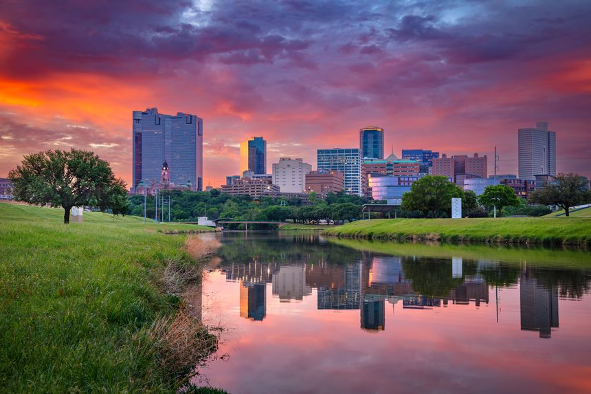 Downtown Fort Worth, Texas, at sunrise. The cityscape features modern skyscrapers reflecting a fiery orange and purple sky. The Trinity River in the foreground mirrors the skyline and vibrant clouds. Lush green grass and a few trees line the riverbanks, adding a natural element to the urban scene. Prominent buildings rise against the horizon, showcasing the architecture of this Texan city.