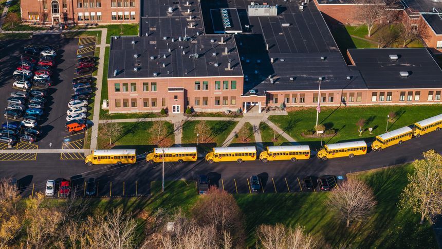 Aerial shot of an American school campus with yellow school buses lined up outside, surrounded by autumn trees and suburban greenery. High quality photo