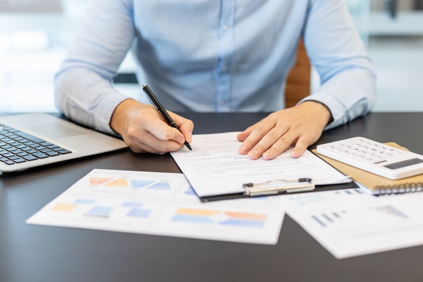 Businessman using a pen to sign a form on a clipboard, reviewing financial documents, graphs, and a calculator on a desk, representing accounting, analysis, and data management
