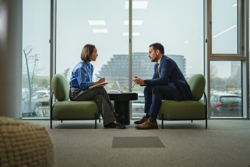 Two diverse professionals in a modern office engaged in a serious interview or meeting, one speaking intently while the other listens and takes notes, emphasizing communication and teamwork
