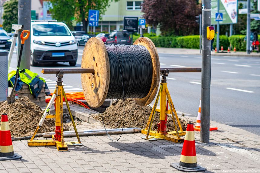 A large wooden cable reel with wound black fiber optic wire mounted on yellow screw jacks on a sidewalk by the street during urban construction and installation works on a sunny day.
