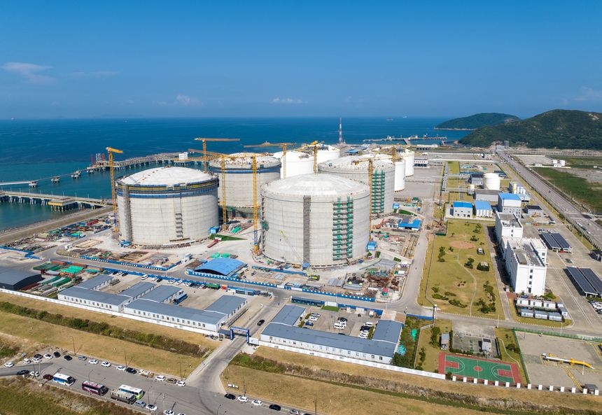 This aerial photograph captures a large liquefied natural gas (LNG) terminal situated along the coast of Zhejiang, China, showcasing its significant storage tanks, docking piers extending into the sea, and extensive industrial infrastructure against a backdrop of blue water and distant hills.A high-altitude view reveals a major LNG terminal in Zhejiang province, highlighting its massive cylindrical storage tanks, intricate network of pipelines and jetties, and the integration of this energy facility with the coastal environment.From above, this image depicts a substantial LNG processing and storage terminal on the Zhejiang coastline, emphasizing the scale of its construction, its proximity to maritime transport, and the surrounding landscape.