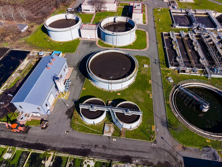 A large water treatment facility is actively processing wastewater. Round tanks hold biosolids as machinery operates. The site is organized, with green grass surrounding the tanks.