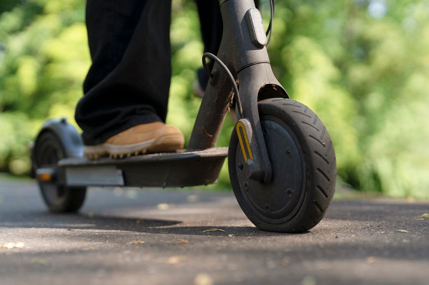 A person stands on an electric scooter on a path. The sun shines down, and trees line the sides of the trail. The scene shows a clear day with no people around.