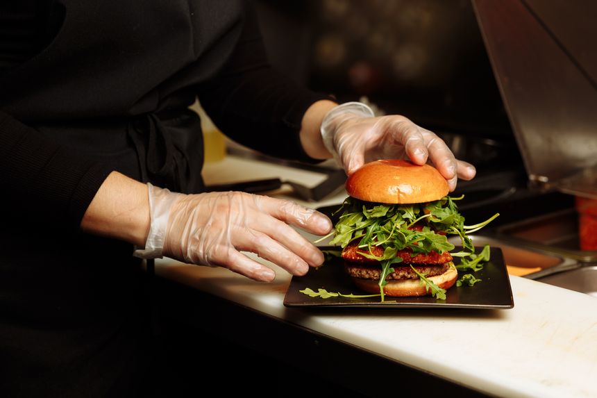 Chef hands in gloves assembling vegan burger with plant based patty and fresh arugula in restaurant kitchen. g.