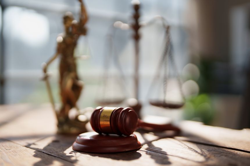 Gavel resting on a wooden desk with a blurred Lady Justice statue and scales of justice in the background, symbolizing law, legal counsel, and justice system
