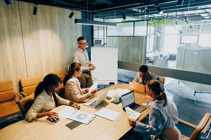 Project planning in modern boardroom, project manager assigning tasks, team updating gantt charts and notes, laptops and printed