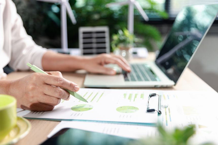 Professional checking ESG investment report with charts and graphs. Background features miniature wind turbines and solar panels, symbolizing renewable energy, green finance, and sustainability.