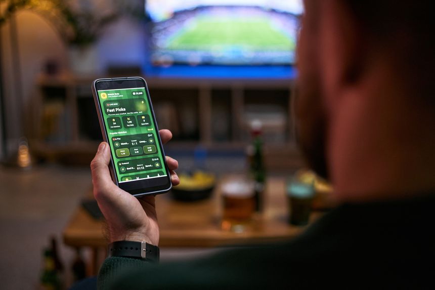 Young adult man holding smartphone, checking sports betting app while watching match with friend, representing male friendship and fan engagement. For gaming, betting, sports marketing