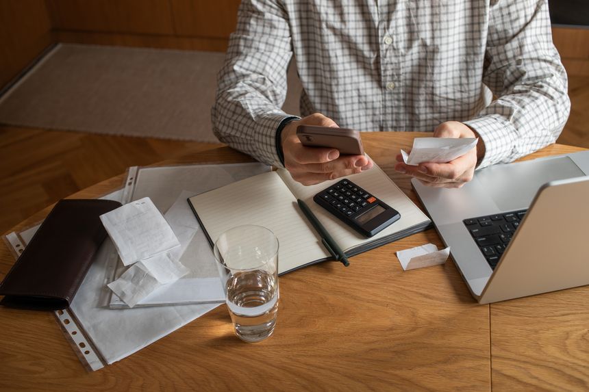 Homeowner man at home kitchen table planning family budget, checking financial unpaid bills for household utilities and mortgage or rent using laptop and phone with internet banking.