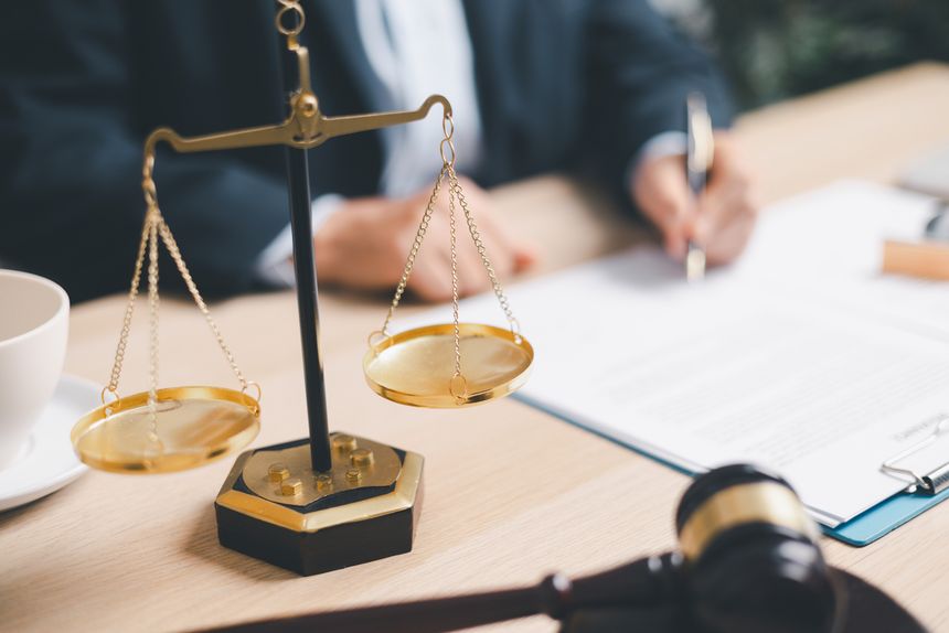 Professional attorney in a suit signing legal documents on a wooden desk with golden scales of justice and a gavel. Concepts of law, arbitration, contract agreement and courtroom authority.
