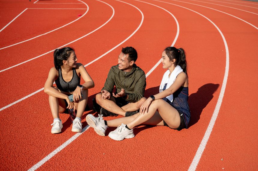 Portrait of a group of cheerful Asian athlete friends in sportswear, enjoying a conversation while resting on a sports track after training and running at a stadium on a sunny day.