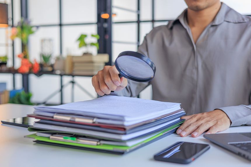 Corporate analyst reviews structured documents and folders with a magnifying glass at a workstation. Concept for financial auditing, detail-oriented research, or risk investigation.