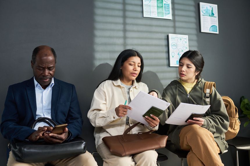 Middle aged Black man using smartphone while two young adult Hispanic women reviewing documents together in visa center waiting area