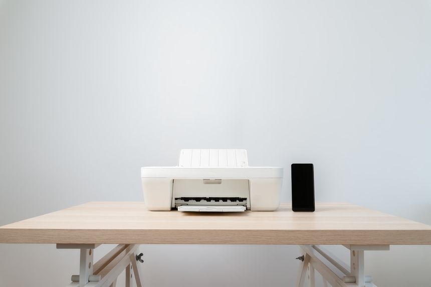 Minimal workspace featuring a white printer and a black smartphone on a light wooden desk against a neutral wall, evoking calm, simplicity, technology, and small home office or studio setup.