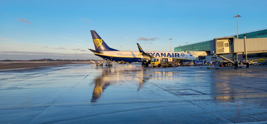 Stansted Airport UK November 12 2025: A Ryanair aircraft is docked at a modern airport gate on a wet tarmac, reflecting the clear blue sky. Ground crew and service vehicles prepare the plane for boarding.Stansted Airport UK 12 November 2025: